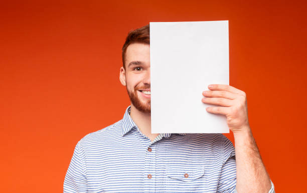 Young Man Holds Blank Paper Against Vibrant Orange Background With a Smile in a Studio Setting