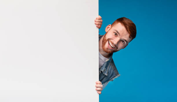 istock Cheerful young guy looking out of white blank placard 2196431679