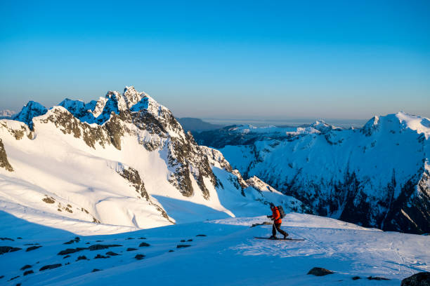 Backcountry skier climbs mountain above valley and ranges at dawn, Tantalus Ranges