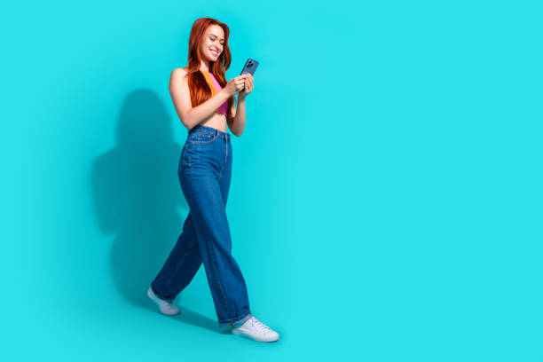 istock Young woman with red hair smiling while using a smartphone against a teal background, wearing casual summer outfit with flared jeans 2196442306