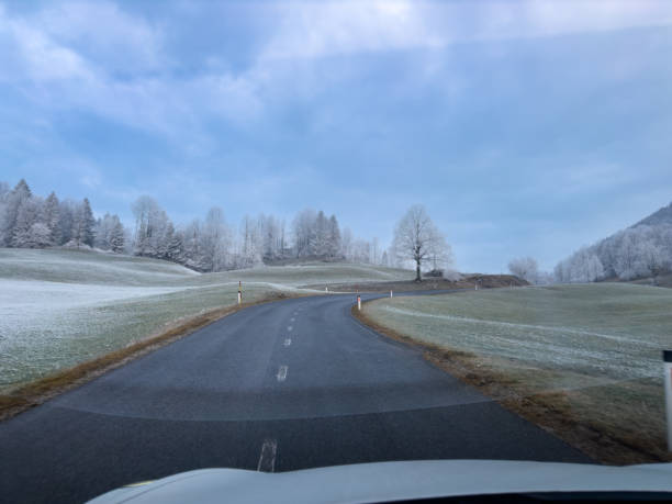 View from a car driving along a winding road through a frosty countryside, with snow-dusted fields, bare trees, and a serene blue sky creating a peaceful winter scene.