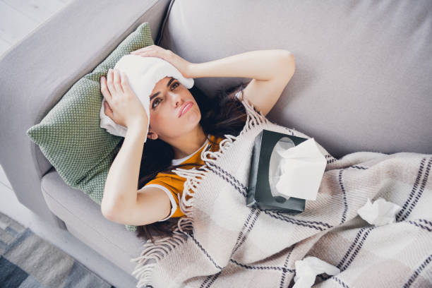 istock Young woman resting on sofa with towel on forehead, wrapped in blanket, feeling unwell 2196448414
