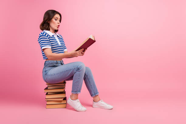 istock Young woman reading a book on a stack of books with a surprised expression against a pink background 2196450867
