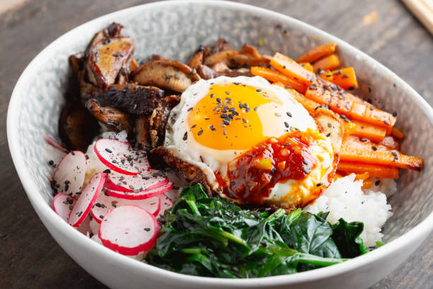 Korean rice dish Bi Bim Bap: a bowl with rice, topped with various vegetables such as fried shiitake mushrooms, spinach, carrots and radish, with a fried egg on wooden background, close-up