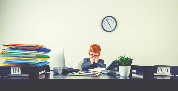 Humorous image of stressed and upset young boy working on a laptop computer at a desk. He is wearing a suit and is looking overworked. There is a phone on his desk along with a computer an inbox, outbox, digital tablet and adding machine