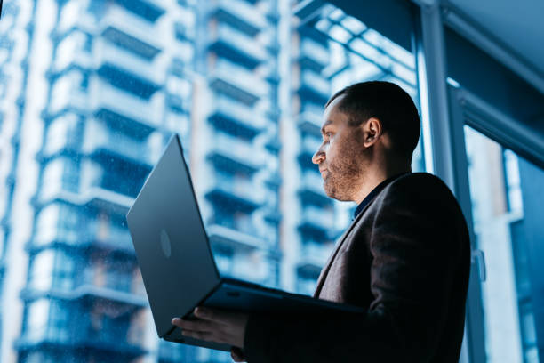 istock A businessman in a modern corporate office holds a laptop, brainstorming ideas to solve a task against a cityscape background. 2196456697