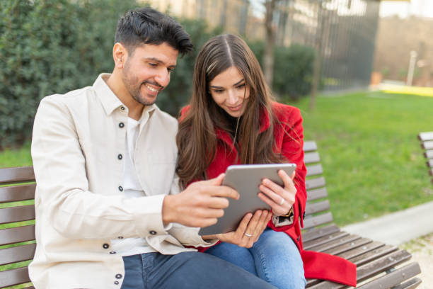 istock Young couple using tablet sitting on bench in a park 2196469517