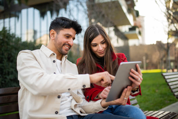 istock Young couple using tablet sitting on bench in city park 2196470372