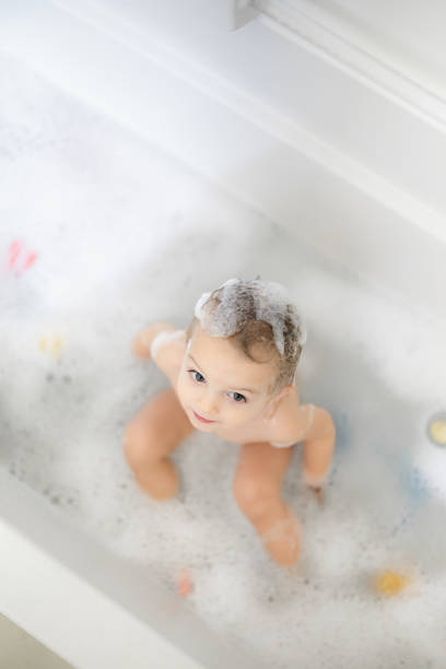 A charming toddler with soap suds on their head enjoys a pleasant bath in a white bathtub. Colorful bath toys and bubbly foam create a joyful and playful atmosphere during bath time.