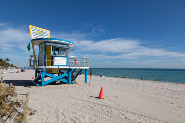 A vibrant blue and yellow lifeguard tower stands proudly on the pristine sands of Hollywood Beach, Florida. Under the morning sun, beachgoers relax by the turquoise waters, while the tower serves as a symbol of safety and coastal charm.