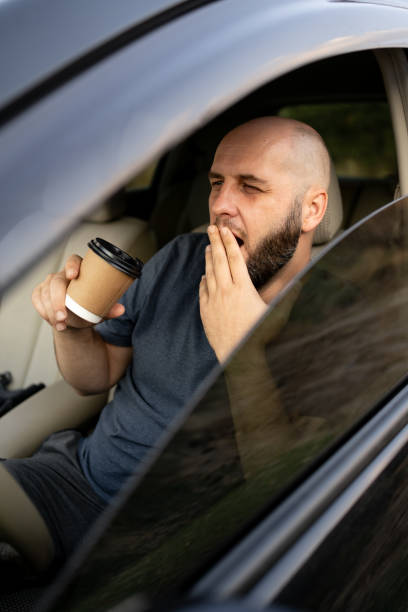 istock Yawning bald bearded man sitting in a car drinking takeaway coffee to avoid falling asleep while driving 2196480767