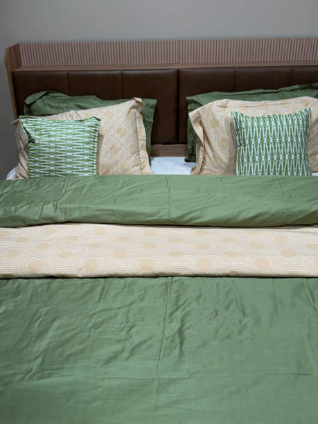 Stock photo showing close-up view of a cosy double bed, with colour-coordinated, fresh bedding in green and beige, plain and patterned textiles. Home soft furnishings and bedroom interior design concept. close-up image of double bed with padded, pleather covered headboard and integrated wooden shelf, co-ordinated brown and green bedding, pillow and cushion stack, white bed sheet, folded duvet, focus on foreground, elevated view, soft home furnishing
