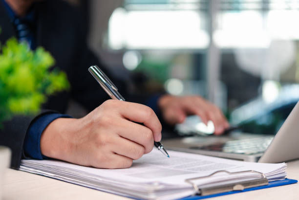 istock Businessman signing documents working on laptop in office environment. Close-up view of executive hand writing with pen on paper documents beside computer workspace. 2196495583