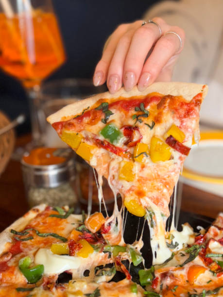istock Close-up image of unrecognisable woman holding thin, crispy crust mixed bell pepper pizza, slice being raised from plate, displaying manicure nail art, glass of Spritz Veneziano, restaurant setting, focus on foreground 2196498999