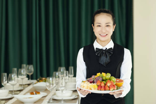 A woman wearing a blue blouse and apron slicing a watermelon on a table, with kitchenware in the foreground, displaying focus and precision in meal preparation in a kitchen setting.