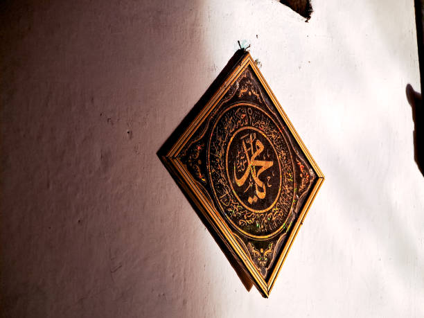 A framed calligraphic artwork featuring the name of the Prophet Muhammad (SAW) in Arabic, hanging on a wall within a mosque