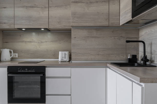 istock Modern built-in kitchen in minimalist style. kitchen unit with wooden upper and beige lower cabinets. built-in oven, microwave. white electric kettle, toaster. black sink, faucet. kitchen backsplash 2196519158