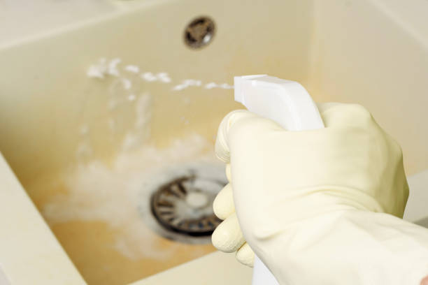 A hand in a rubber glove sprays an alkaline cleaning solution onto a dirty kitchen sink. Foam begins to break down plaque and stains.