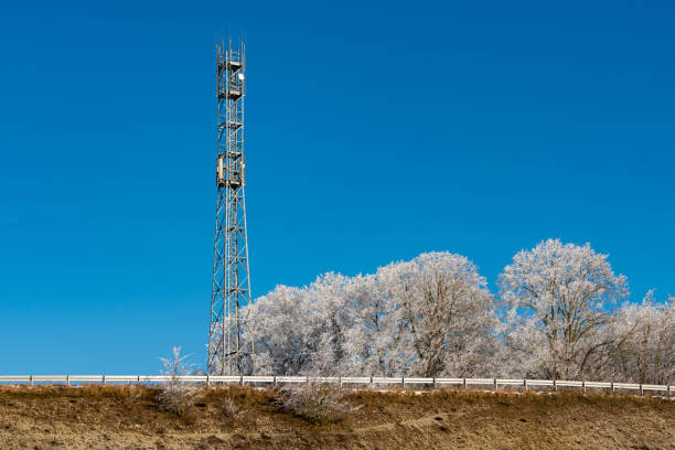 A communication tower rises prominently beside a frost-covered landscape. The bright blue sky creates a striking contrast with the white trees nearby, emphasizing the wintry scene.