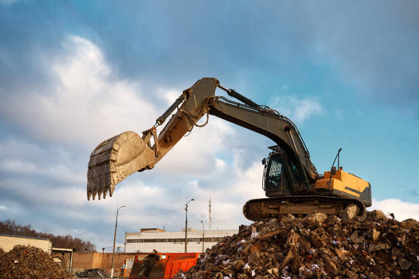 Building excavator with large bucket works on pile of concrete waste at factory demolition site against grey cloudy sky at sunset
