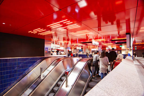 A subway station interior with glossy red ceiling panels reflecting the bright lights, escalators leading to the platform, and people descending with a blue-tiled wall adding contrast.