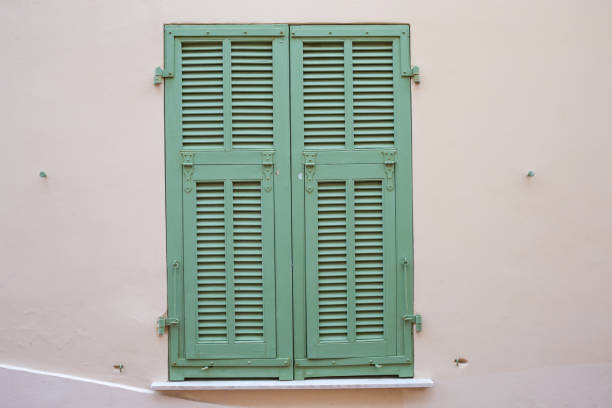 Window with closed wooden shutters painted green installed in beige wall outdoors in overcast day