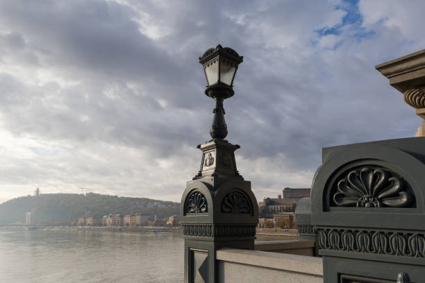 Decorative street lamp installed on the Szechenyi Chain Bridge of the 19th century against the River Danube, western hilly riverbank and cloudy sky in autumn overcast morning, Budapest