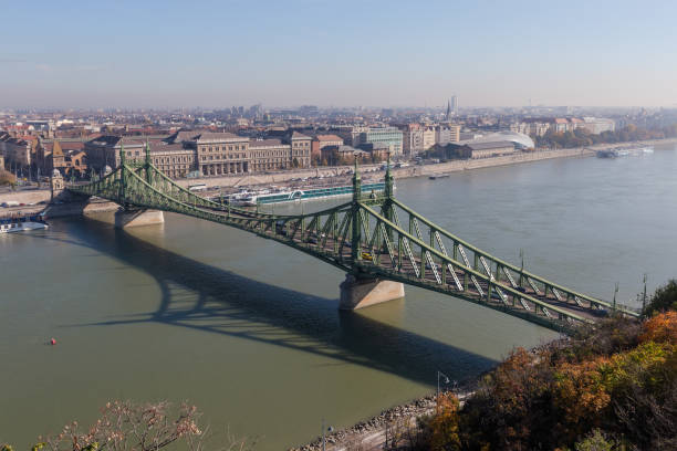 Budapest, Hungary - April 2025 A wide-angle view of the iconic Danube River in Budapest, Hungary, viewed from Buda across to the banks of Pest. Several boats, including a cruise ship and smaller vessels, navigate the gray waters under a cloudy sky. The scene captures the historic architecture against a backdrop of urban buildings and distant green hills, evoking a sense of timeless European cityscape. No people are present. This image suits commercial uses in travel brochures, tourism websites, urban planning articles, historical documentaries, and media content highlighting Budapest's landmarks for authors and publishers.A wide-angle view of the iconic Széchenyi Chain Bridge crossing the Danube River in Budapest, Hungary, with the distinctive Hungarian Parliament Building visible on the riverbank. Several boats, including a cruise ship and smaller vessels, navigate the gray waters under a cloudy sky. The scene captures the historic architecture against a backdrop of urban buildings and distant green hills, evoking a sense of timeless European cityscape. No people are present. This image suits commercial uses in travel brochures, tourism websites, urban planning articles, historical documentaries, and media content highlighting Budapest's landmarks for authors and publishers.
