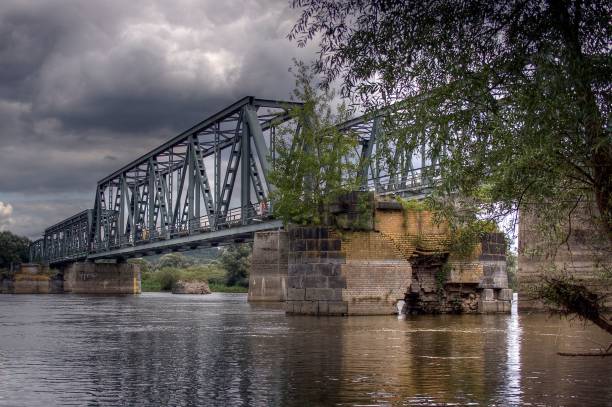 A steel bridge over a calm river with a cloudy sky and surrounding greenery