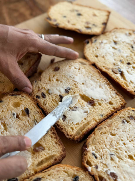 istock Close-up image of unrecognisable person in kitchen setting, buttering panettone, Italian bread loaf on wooden chopping board, freshly sliced fruit cake, spreading butter with knife, wood grain kitchen counter, focus on foreground 2196541624