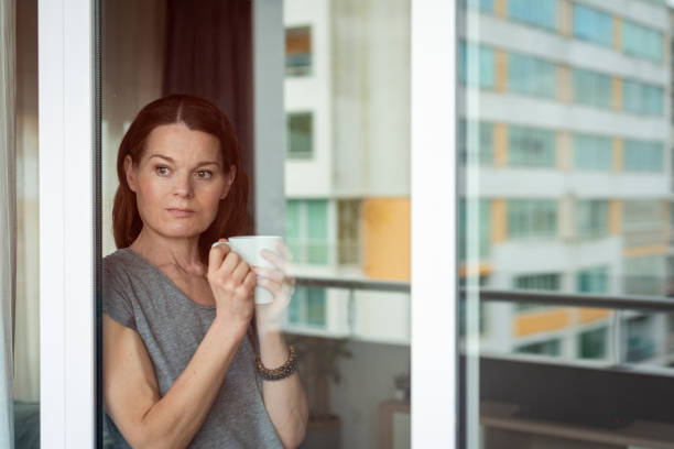istock woman standing by a window holding a mug, reflections 2196547874