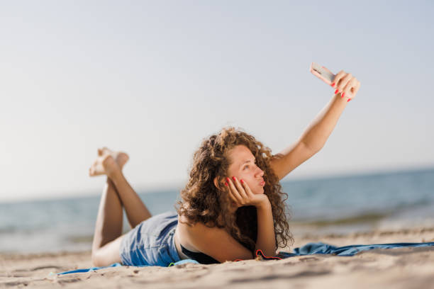 istock Young woman taking a selfie at the beach while lying on a towel on a sunny day 2196561433