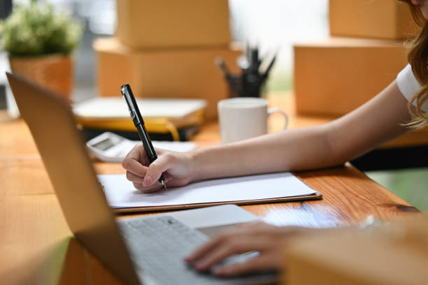 istock Close-up of a small business owner writing in a notebook while using a laptop, surrounded by packages ready for shipment 2196573032