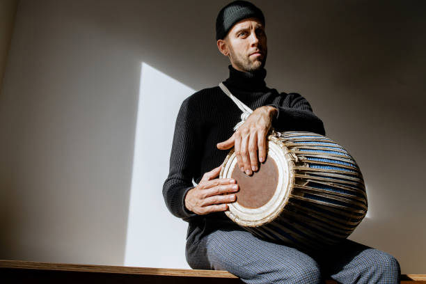 istock Man in black clothes and cap sitting and having joy while playing mridangam, an ancient percussion musical instrument originating from the Indian subcontinent in sunny day inside. Copy space 2196581526