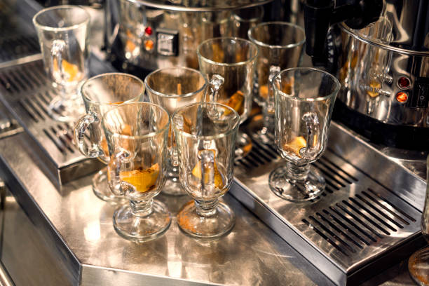 Empty irish coffee glasses are waiting to be filled, placed on a stainless steel counter top near a coffee machine, with lemon and cinnamon inside