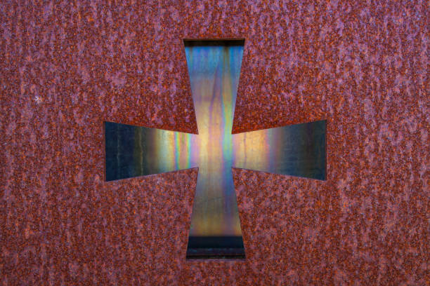 A metallic cross with a textured, iridescent surface is centrally framed against a dark, rough background with a subtle reddish hue.