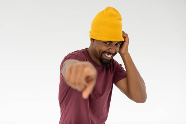 An excited man wearing a bright yellow beanie points playfully at the camera, exuding enthusiasm and a carefree spirit against a minimalist white backdrop.