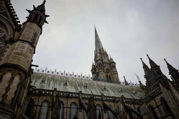 An upward view of the intricate Gothic architecture of St. Colman's Cathedral in Cobh, Ireland. The image highlights the cathedral's towering spire and ornate details against a cloudy sky
