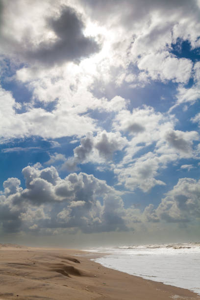 A vast sandy beach stretches towards a turbulent ocean under a dramatic sky filled with fluffy white clouds and patches of blue.