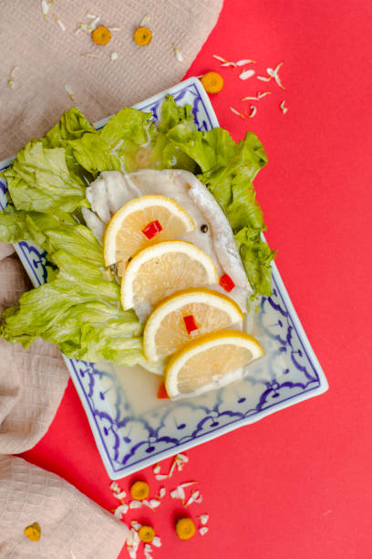 Top view of steamed fish fillet with lemon slices, chili, and fresh lettuce served on a traditional blue-patterned plate against a vibrant red background, Thai seafood cuisine concept
