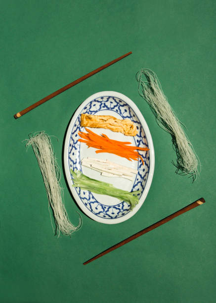 This image showcases a traditional Thai rice noodle dish with colorful strands of orange, white, and green noodles elegantly presented on a decorative ceramic plate. Surrounding the plate are dried rice noodles and wooden chopsticks, arranged on a bold green background that enhances the vibrancy of the composition. Ideal for use in Thai cuisine promotions, restaurant branding, or cultural food photography, this image highlights the artistry and authenticity of Thai culinary traditions.
