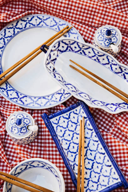 This image features a set of traditional Thai ceramic tableware with elegant blue patterns, carefully arranged with wooden chopsticks on a red checkered fabric background. The collection includes plates, bowls, and tea cups, reflecting the cultural richness of Thai and Asian dining traditions. Ideal for use in restaurant branding, food photography, or cultural promotions, this image captures the beauty and authenticity of traditional table settings.