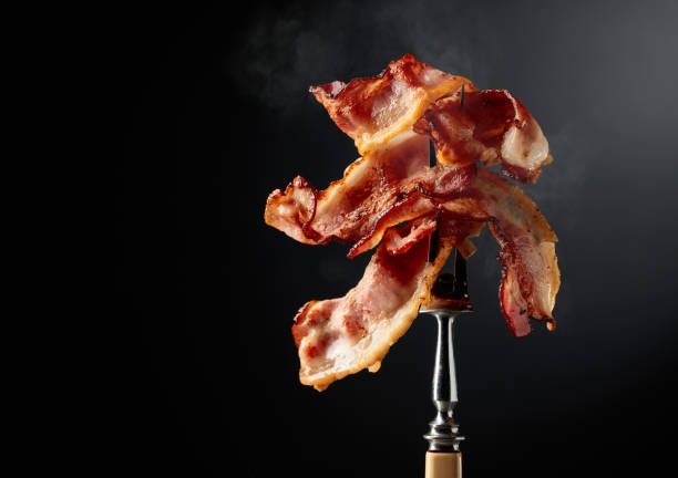 Fried bacon on a fork. Fried bacon on a fork against a black background. Copy space. cooked bacon slice stock pictures, royalty-free photos & images