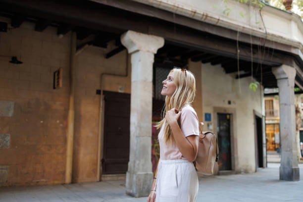 Blonde woman tourist carrying a backpack strolls through a charming european city, admiring the stunning architecture and historical buildings