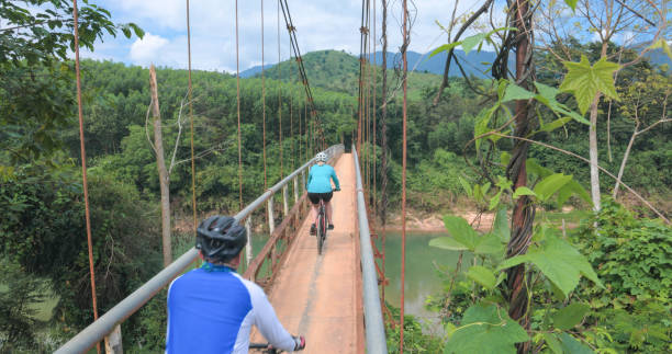 Bicyclist and guide ride suspension bridge over river, tropical rainforest and mountains distant
