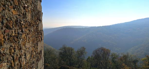 A breathtaking view of rolling mountains covered in dense forests, partially veiled in a soft mist, as seen from the edge of an ancient stone structure. The rugged texture of the stone wall contrasts with the vast, serene landscape, creating a timeless and mysterious atmosphere.