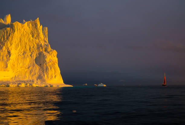 red sailboat sailing among floating yellow icebergs hit by setting sun in Disko Bay during polar summer midnight solar season. Ilulissat, Greenland