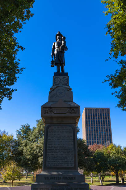 The Volunteer Firefighter Memorial, located on the grounds of the Texas State Capitol in Austin, Texas, honors the bravery and sacrifice of volunteer firefighters. The bronze statue depicts a firefighter carrying a child in one arm while holding a lantern in the other, symbolizing rescue and dedication to service. The monument, framed against a clear blue sky and surrounded by lush greenery, serves as a tribute to those who risk their lives to protect their communities.