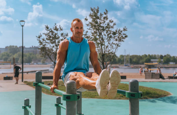 A man in a blue tank top performs an L-sit on parallel bars in a park, showcasing impressive core strength and focus, amidst a sunny day ambiance.