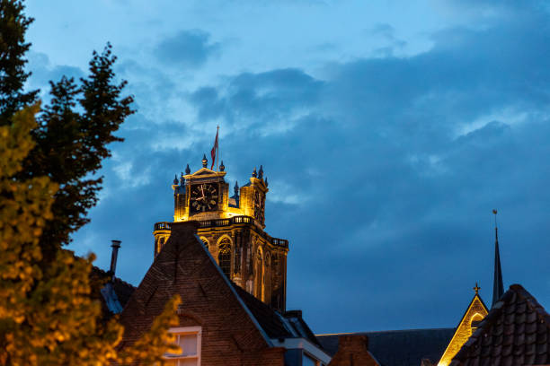 An illuminated historic clock tower rises above the rooftops at dusk, with blue sky in the background. Warm lights highlight the architectural details, creating contrast with the evening sky.